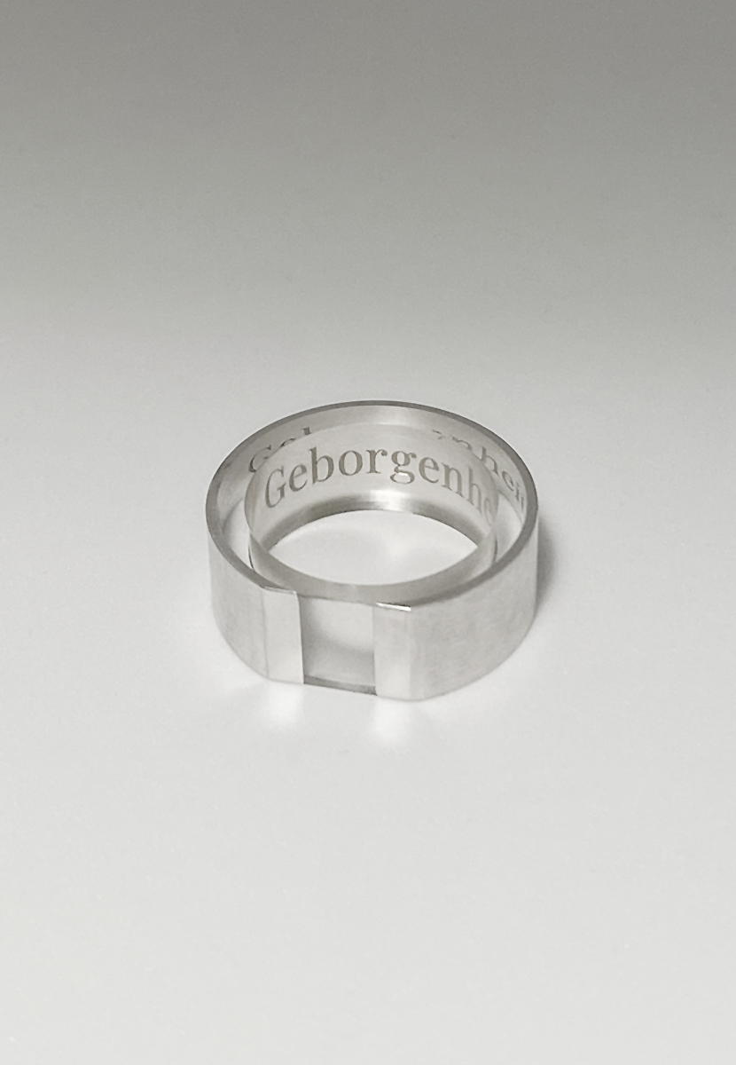 Ein schlichter, silberner Ring mit einer doppelten Bandführung und der eingravierten Aufschrift 'Geborgenheit' in klassischer Typografie, der auf einer hellen Unterlage präsentiert wird, um eine Atmosphäre von Reinheit und Einfachheit auszustrahlen.